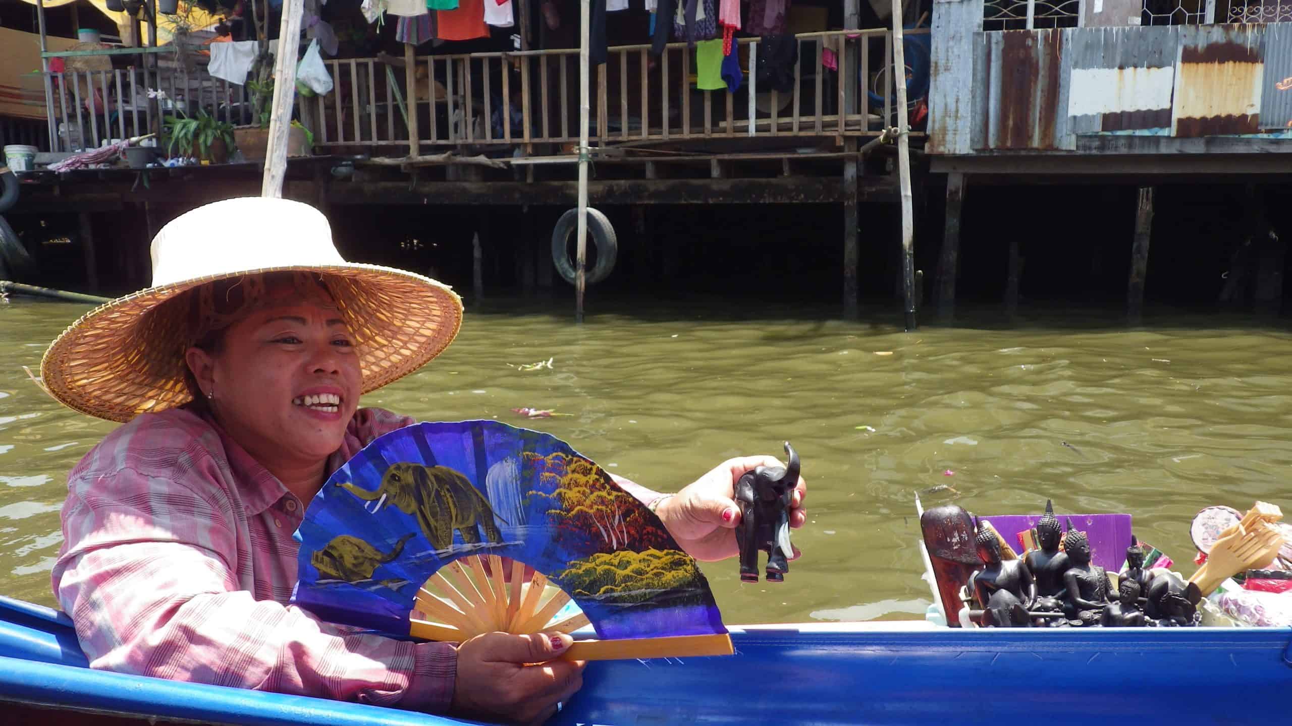 A Thai woman in a straw hat sits in a long boat at a river market near Bangkok selling hand fans and elephant statuettes. 