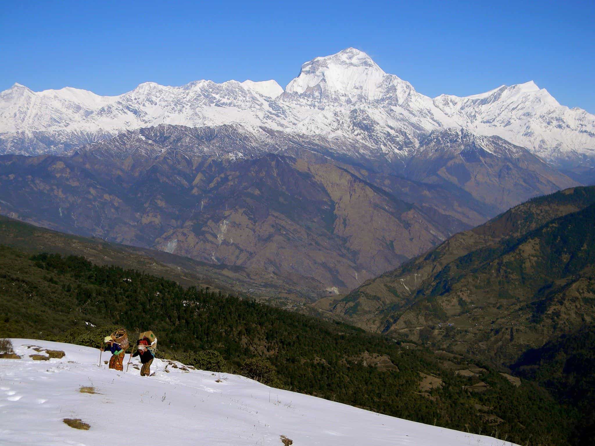 Two Nepalese women climb a snowy mountain carrying heavy loads.
