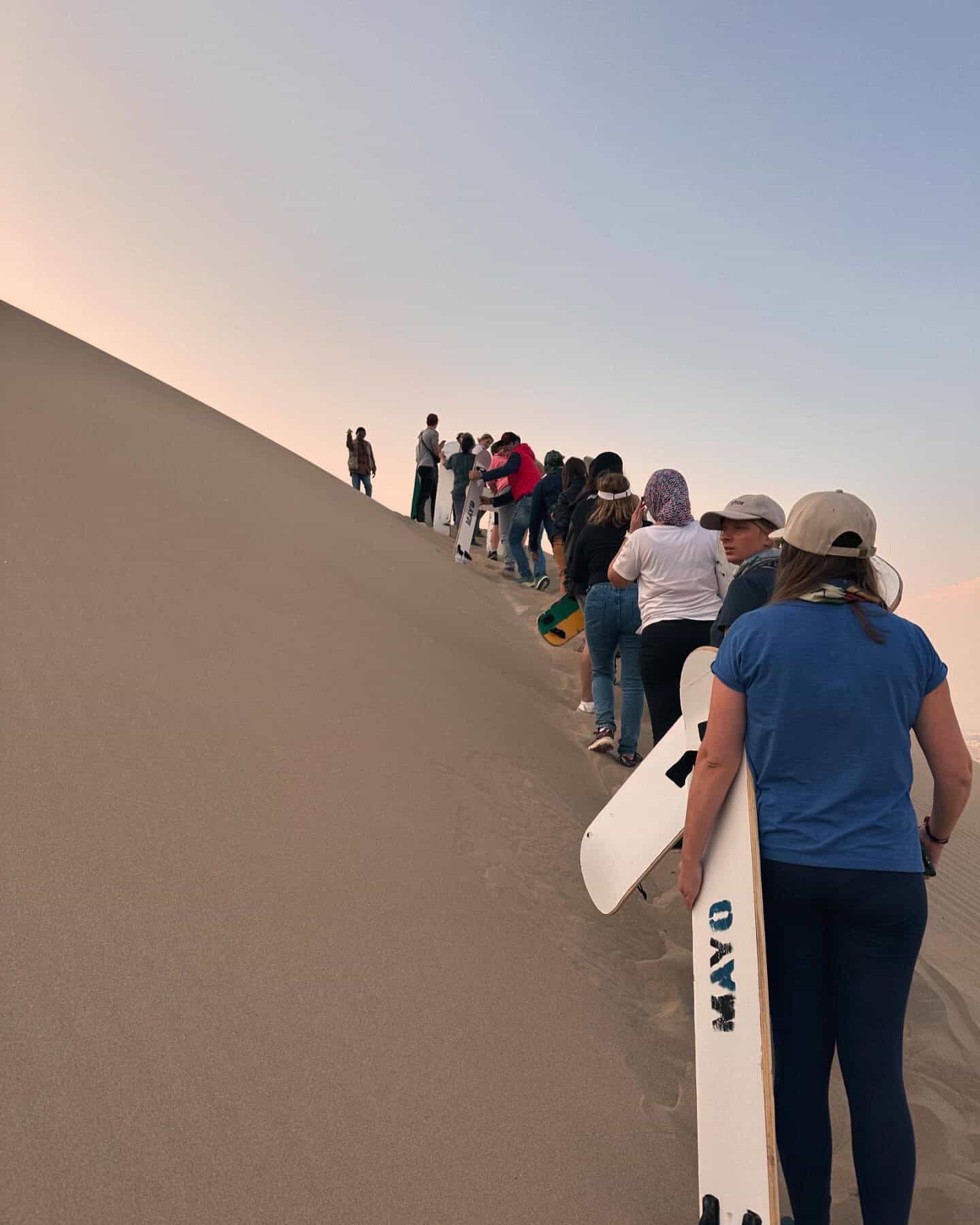 A line of tourists holding sandboards climb a desert dune on a Huacachina Oasis tour.