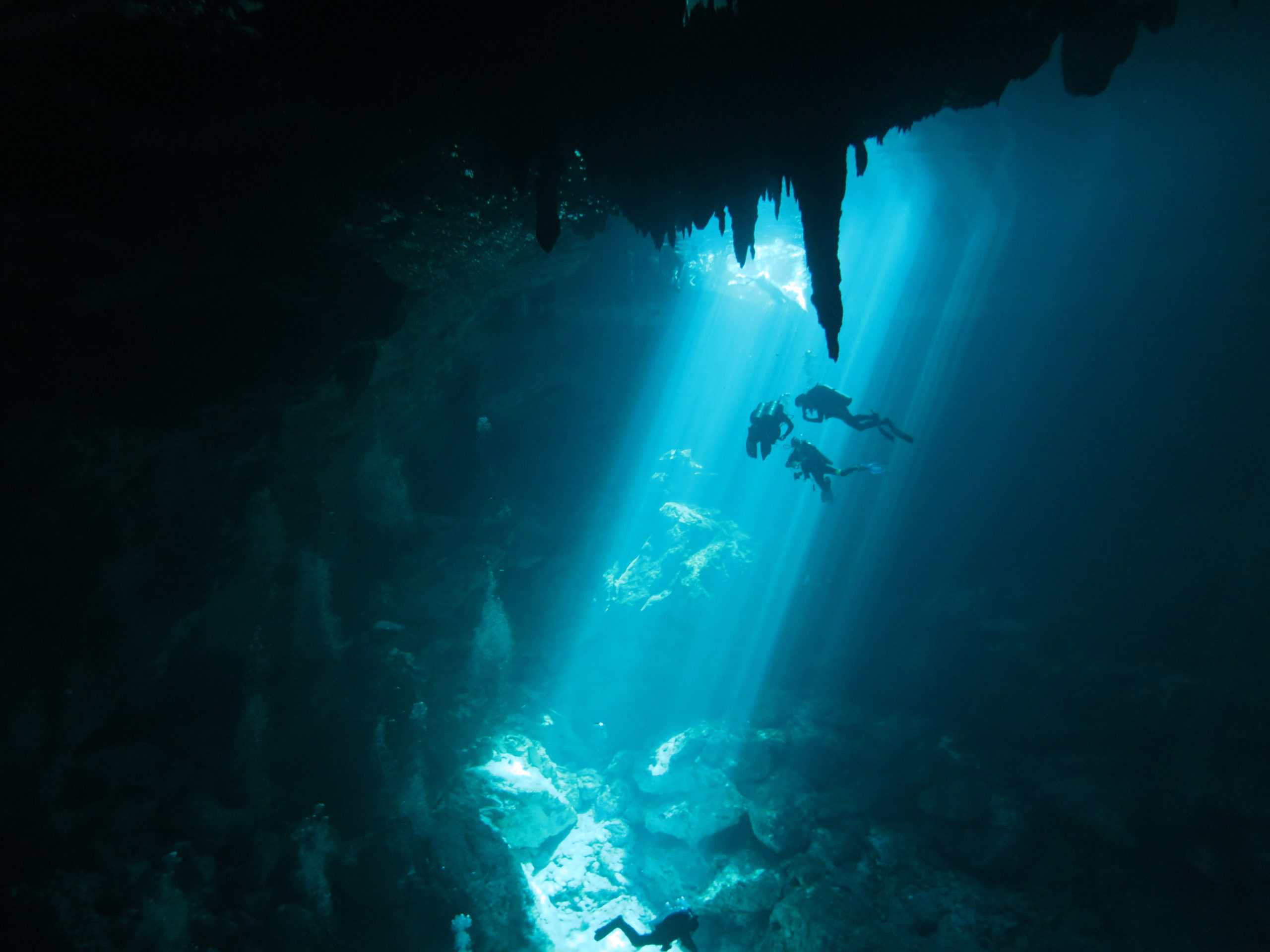Three scuba divers descend into a cenote in Mexico called The Pit while a stream of natural light through the water guides their way.
