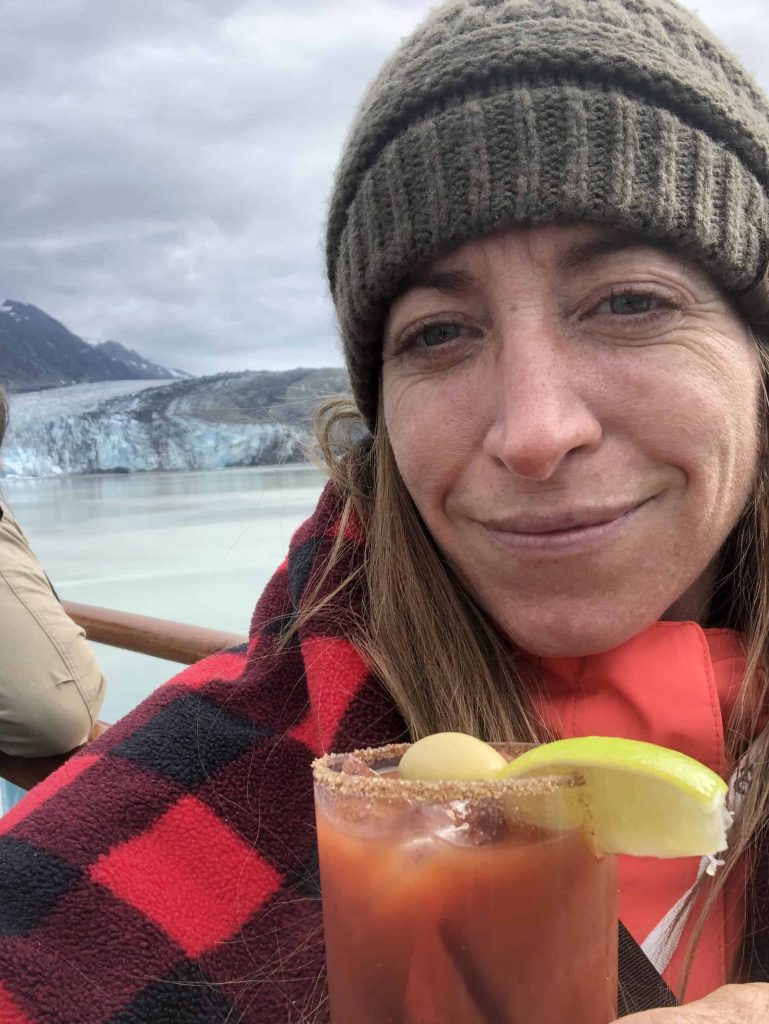 Woman enjoys a bloody mary aboard her Alaskan Cruise with a glacier in the background.