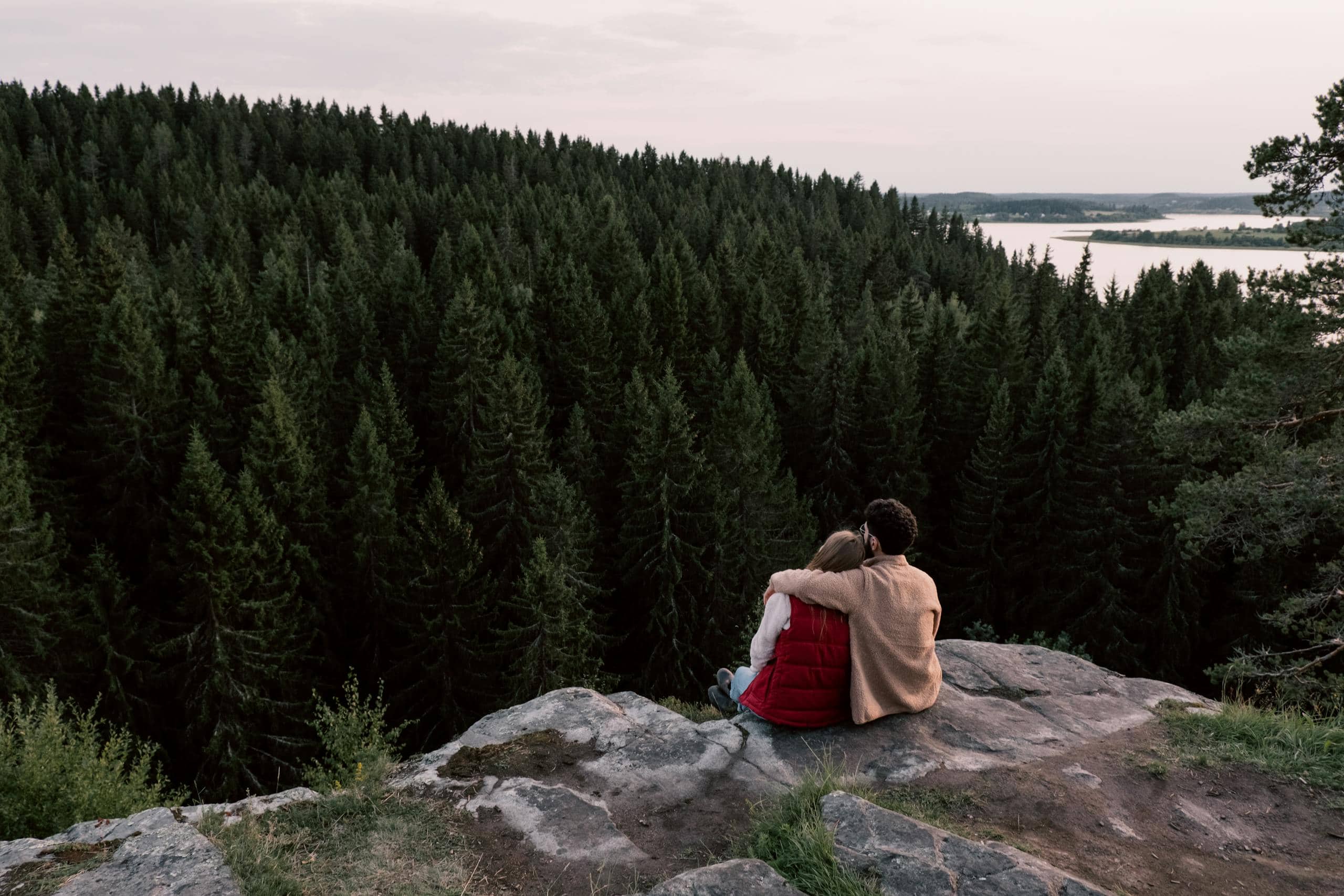 A couple sitting on a cliff, embracing while overlooking a dense forest and lake in Canada.