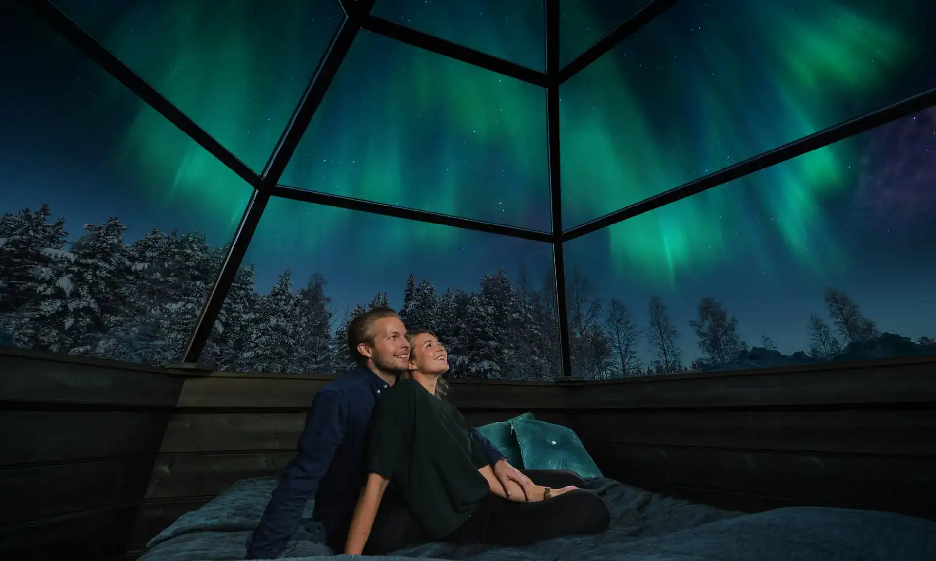 Couple sits inside a cozy glass igloo in Finland as the Norther Lights shine above them.