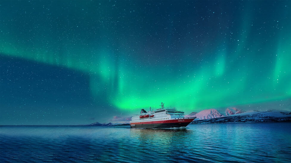A cruise liner sails at night with the Northern Lights dancing in the sky above.