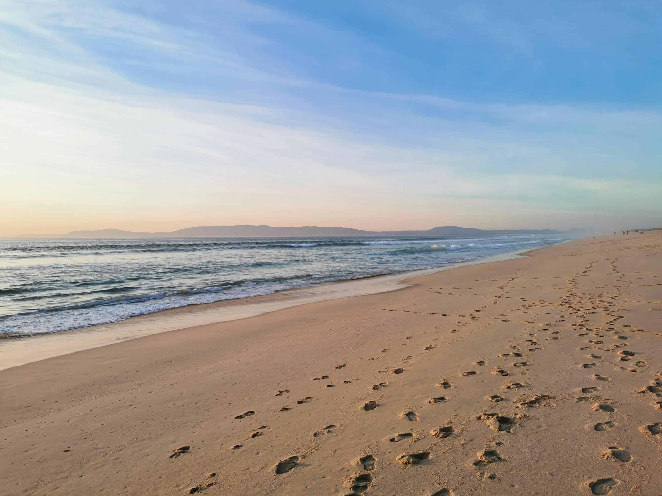 Peaceful sandy shore with footprints and gentle waves at sunset in Comporta, Portugal.