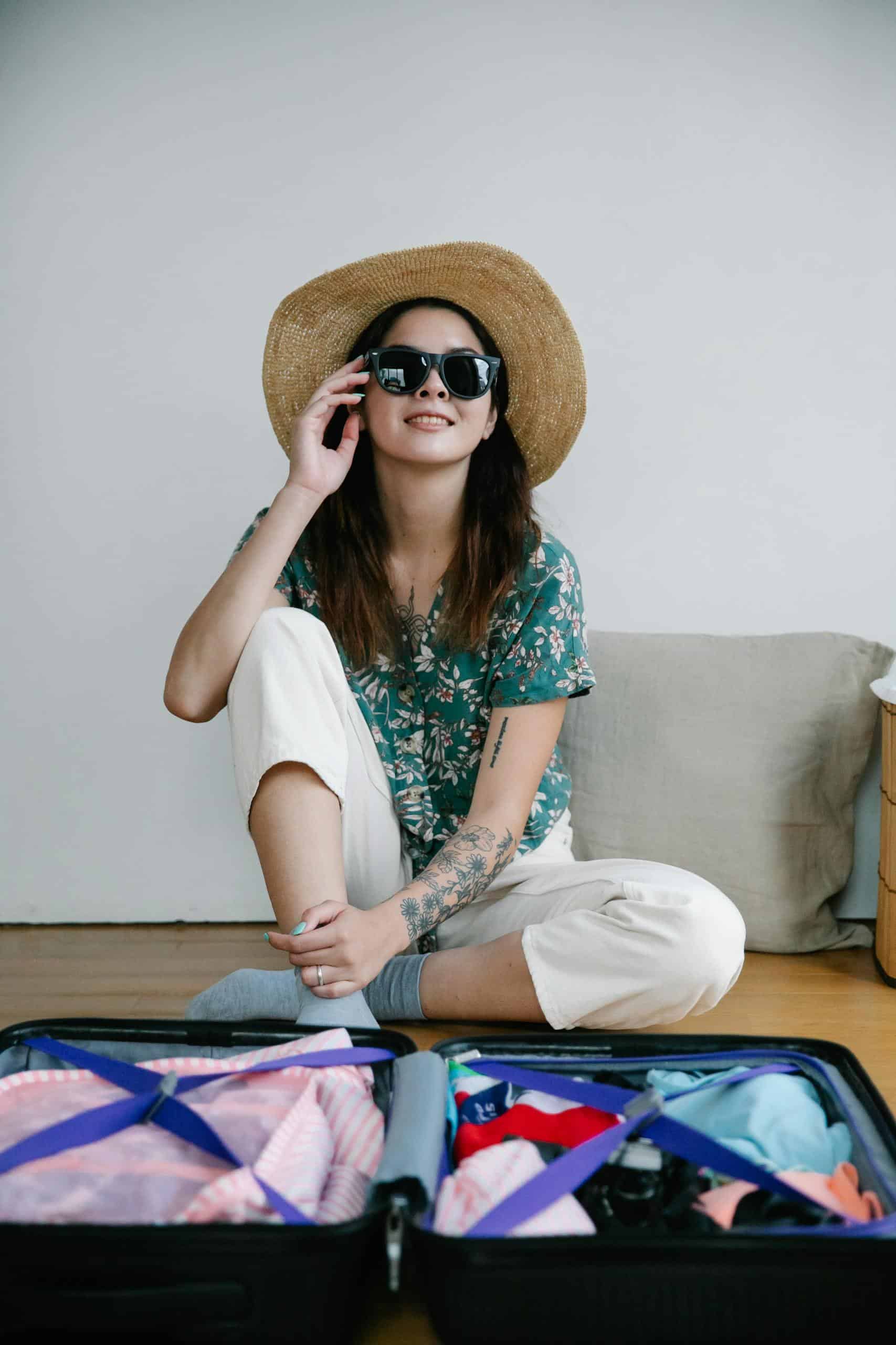 Woman in a hat and sunglasses sitting by a packed suitcase, ready for travel.