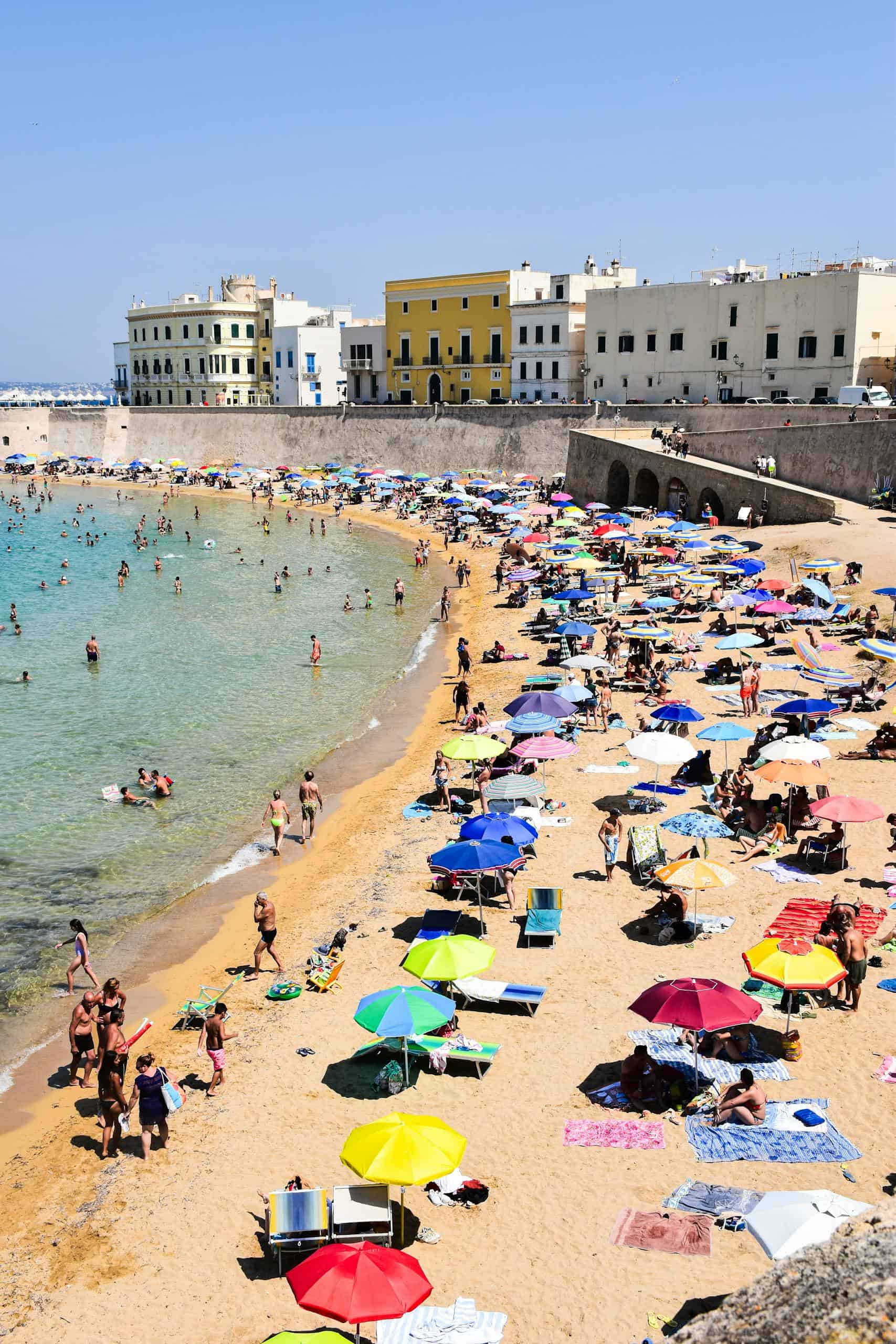 Crowded summer beach scene in Gallipoli, Apulia, Italy, with colorful umbrellas and historic buildings.