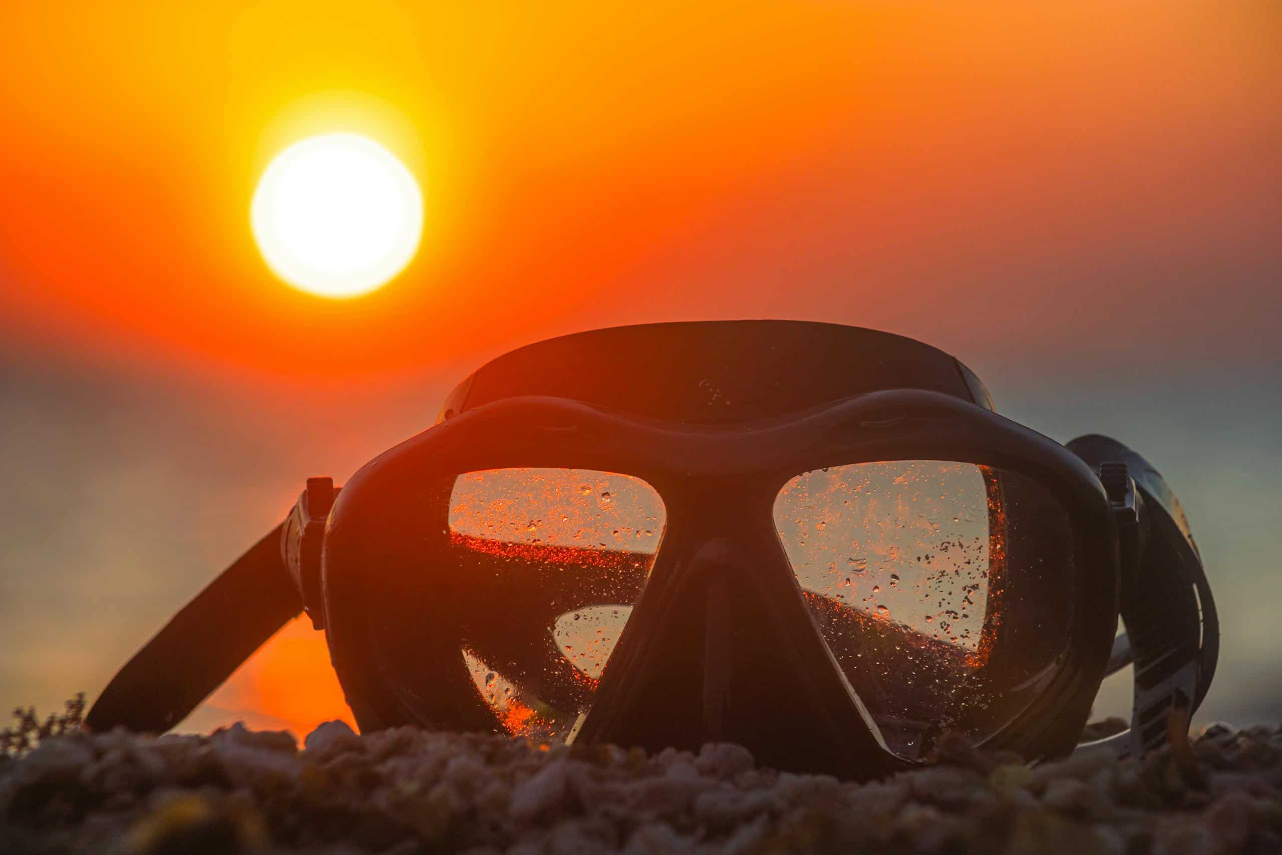 A diving mask rests on a sandy beach with a vibrant sunset reflecting in its lenses.