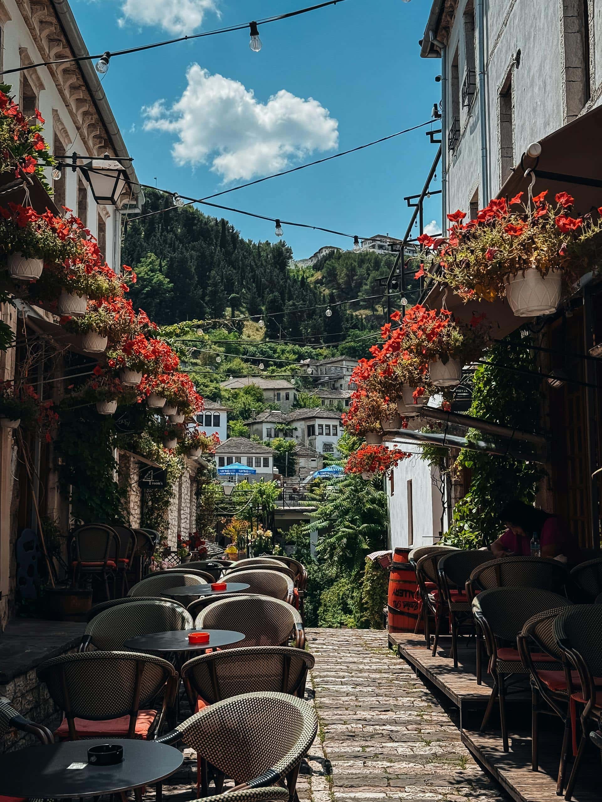 Explore the beautiful historic streets of Gjirokastรซr, Albania featuring vibrant flowers and inviting cafes.
