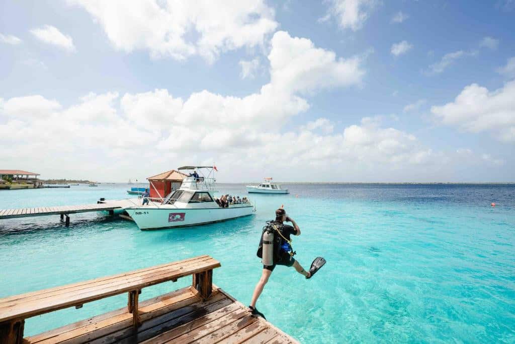 Diver in Bonaire does a giant stride off of a dock with a dive boat floating in the background.
