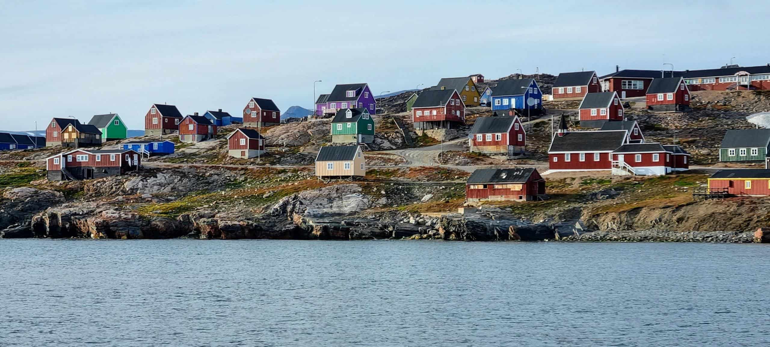View of colorful houses on a rocky shoreline in Ittoqqortoormiit, Greenland, against a clear sky.