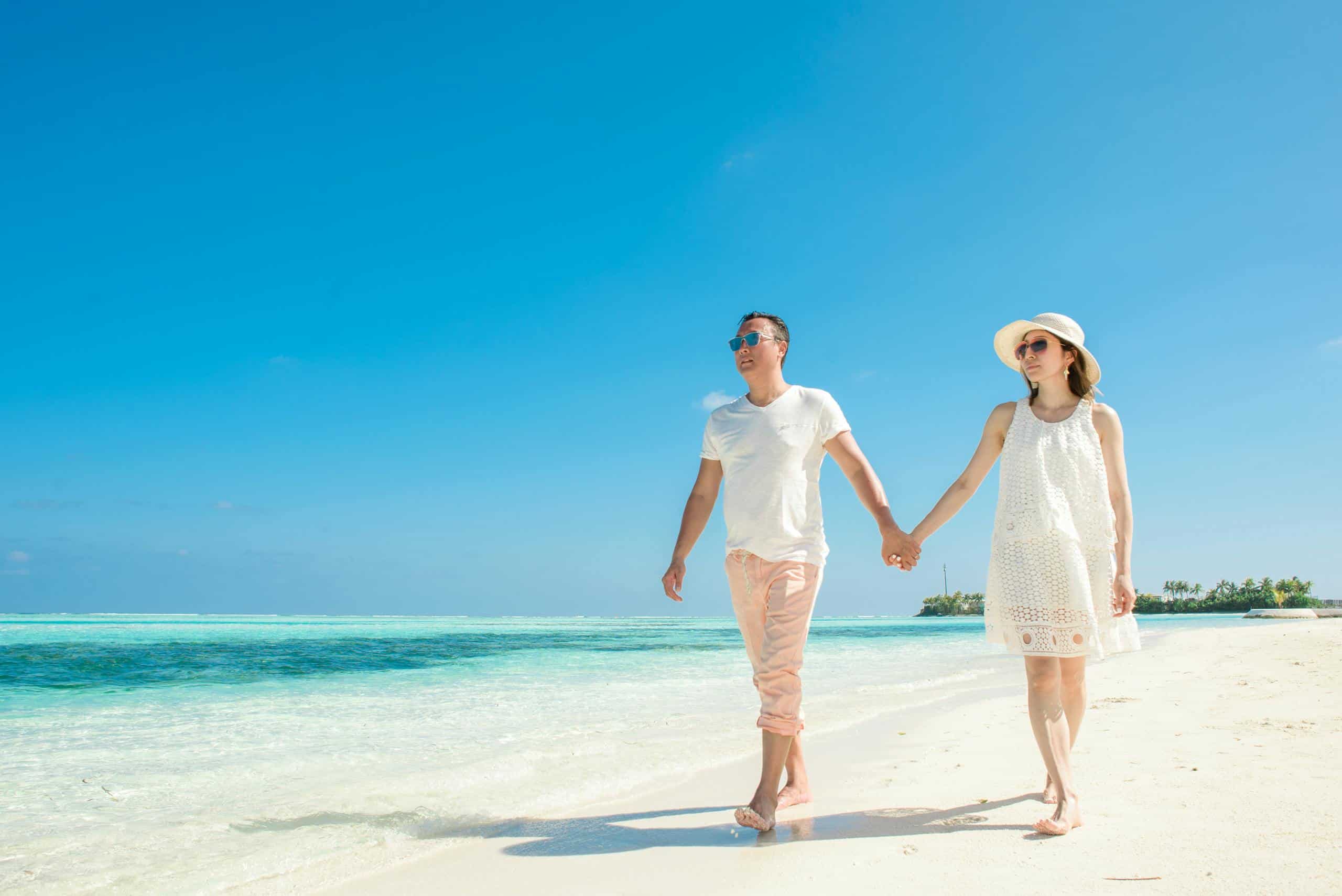 A couple holding hands and walking barefoot on a sandy beach under a clear blue sky.