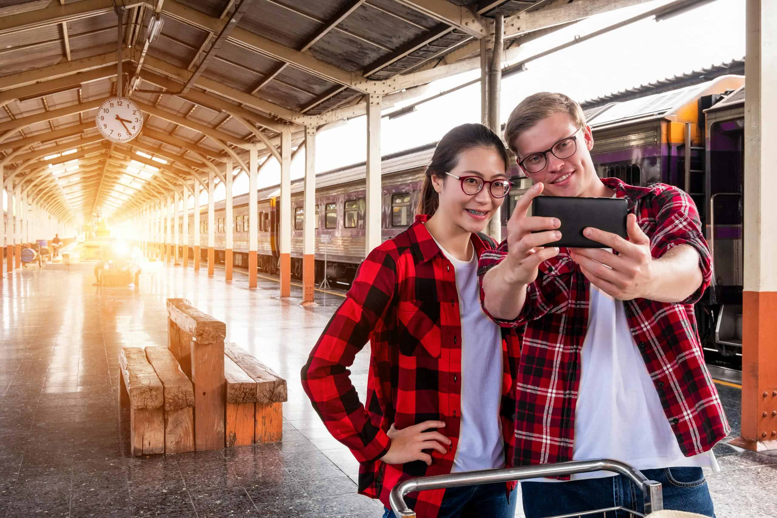 A smiling couple takes a selfie at an urban train station, showcasing travel and fashion.