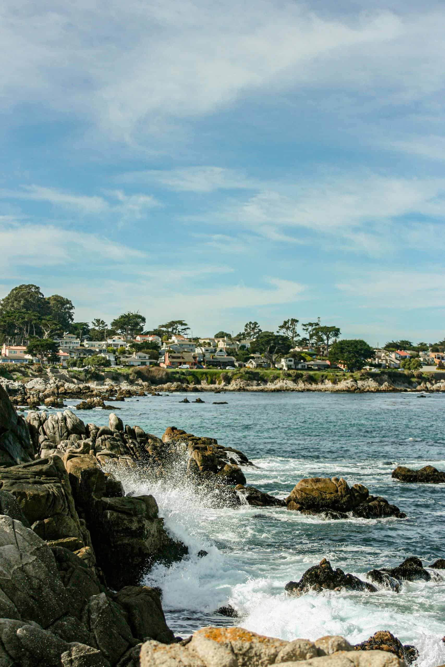 Waves crashing against rocky shore in Carmel-by-the-Sea with distant view of coastal town homes.