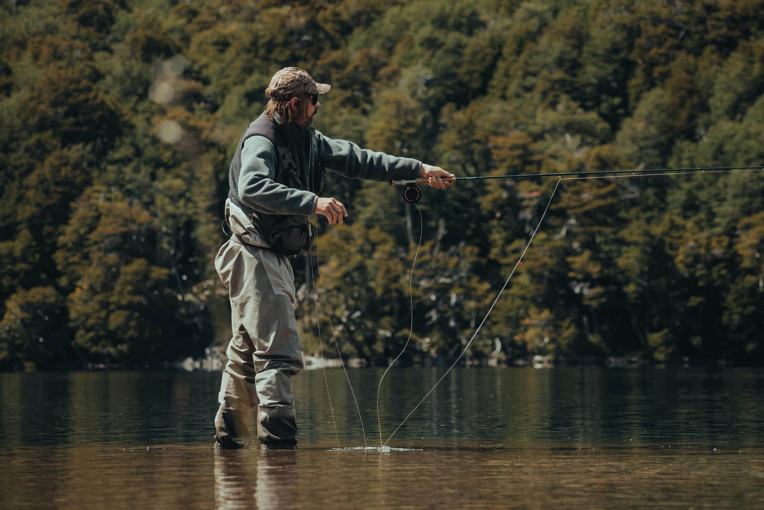 A man in waders fly fishing in a tranquil lake surrounded by lush green forest.