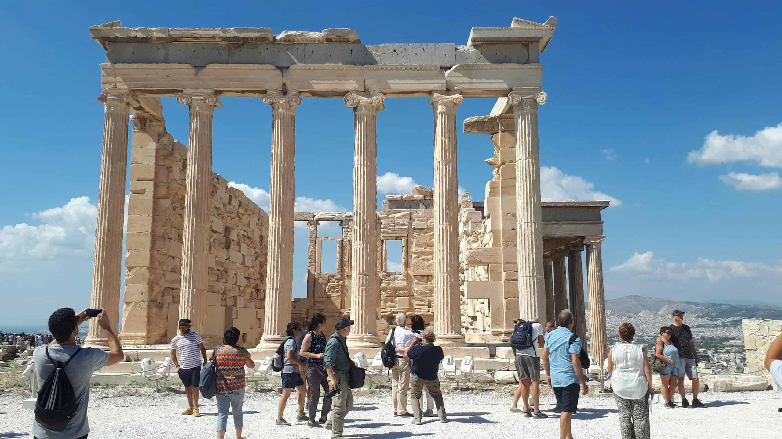 Visitors explore the iconic ruins of the Acropolis under a bright summer sky in Athens, Greece.