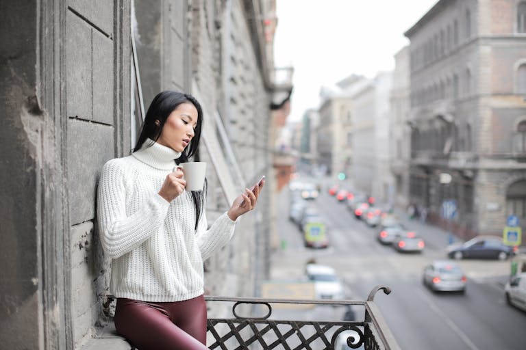 Young woman sipping coffee and using phone on a city balcony.